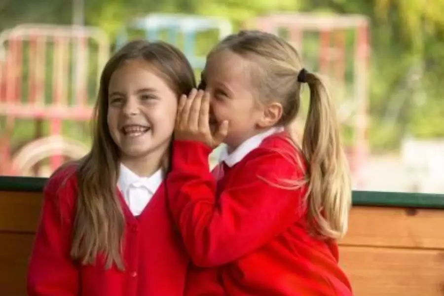 Two happy primary school girls whispering to eachother | Teachers To your Home
